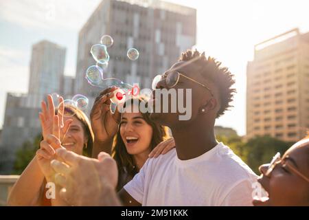Group of multiracial happy friends having fun with soap bubbles in the city - Cheerful young friends smiling with bubbles on sunny day - Hapiness Stock Photo