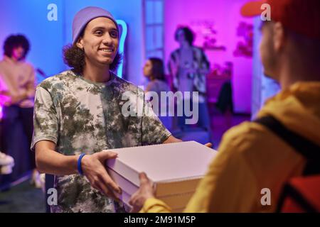 Happy guy looking at courier and taking stack of square cardboard boxes with ordered pizza against his friends enjoying home party Stock Photo