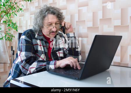 A portrait of an elderly man working at the laptop and phone at home. Technologies, ageing Stock Photo