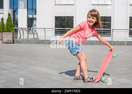An outdoors portrait of a happy little girl with a skateboard on a summer sunny day in the park on a river bank Stock Photo