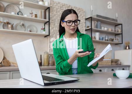 Online business meeting. Young upset woman talking on video call from laptop with tax, bank, creditor. Quarrels, shows a notebook with bills. Stock Photo