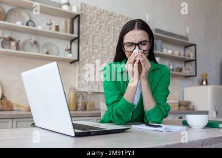 Tired young female freelancer student sitting at home in kitchen with laptop. Works. studies online. sick, feels bad, wipes the runny nose with a napkin. Stock Photo