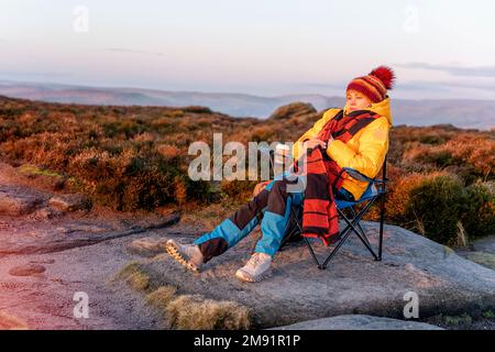 Woman in yellow coat and orange scarf reaching the destination and sitting on beach chairs on top of hill or mountain at sunrise. British cold winter. Stock Photo
