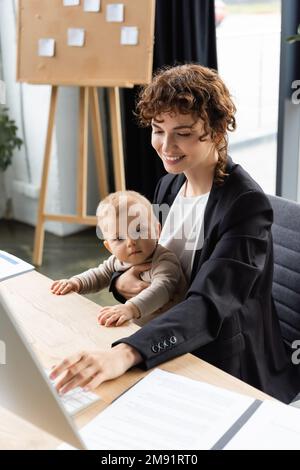 smiling businesswoman working on computer while sitting at workplace with toddler daughter,stock image Stock Photo