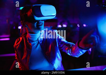 Neon metaverse futuristic concept. elegant middle aged woman in virtual reality goggles exploring metaverse in modern office. Stock Photo