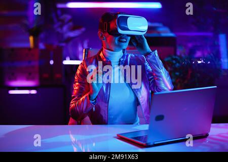 Neon metaverse futuristic concept. happy elegant female in virtual reality goggles designing virtual reality in modern office. Stock Photo