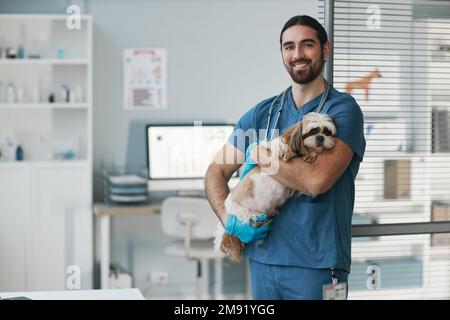 Happy young male veterinarian in blue uniform holding cute yorkshire terrier and looking at camera while standing in medical office Stock Photo