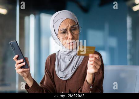 Upset and disappointed woman in hijab inside office trying to make purchase in online store, business woman holding smartphone and bank credit card, received money transfer error. Stock Photo