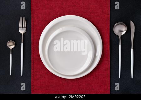 Festive place setting with red napkin. Empty white plates and silver cutlery on black background. Top view. Dining table in restaurant. Card or menu t Stock Photo