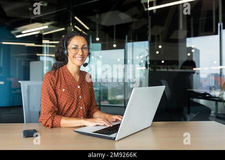 Portrait of Latin American business woman, office worker looking at camera and smiling, using headset and laptop for remote online communication, customer support tech call center worker. Stock Photo