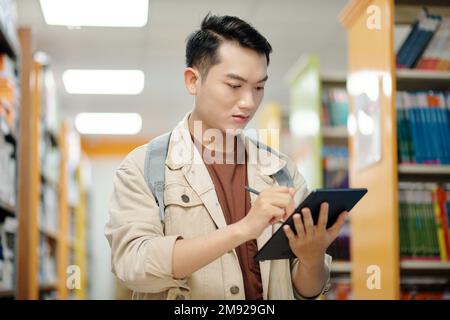 Serious college student standing in library and taking notes on tablet computer Stock Photo