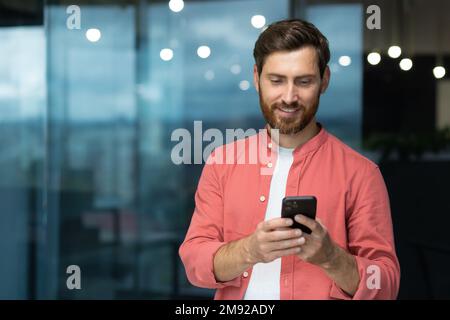 smiling and happy businessman inside the office by the window uses the phone a mature man with a beard looks at the smartphone screen, browses Internet pages, types messages in an online application Stock Photo