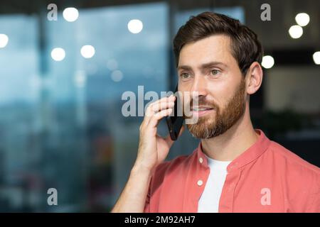 Successful businessman in a red shirt talking on the phone inside the office, a man with a beard at the workplace is talking to a client, a satisfied boss in casual clothes. Stock Photo