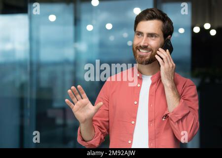 Successful businessman in a red shirt talking on the phone inside the office, a man with a beard at the workplace is talking to a client, a satisfied boss in casual clothes. Stock Photo