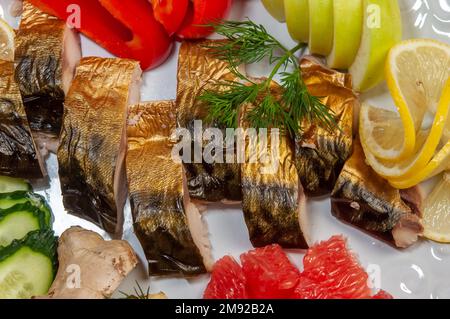 Slices of smoked fish mackerel or scomber on a white dish with lemon, apple, ginger, cucumber, bell pepper, grapefruit, dill, top view. Stock Photo