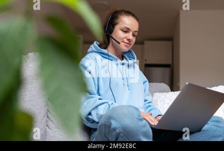 Brunette woman in headset sitting on sofa at home and using laptop working at home remotely e-learning. Stock Photo