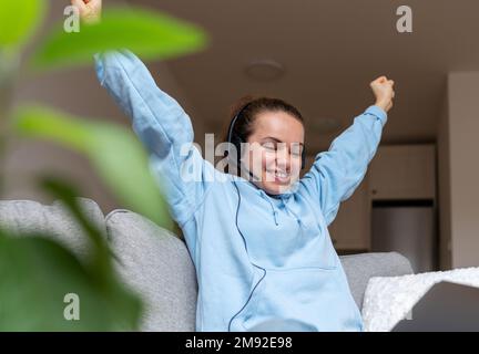 Young woman with headset and arms raised happy looking at laptop while sitting on sofa at home. Stock Photo