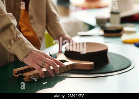 Side view of craftswoman hand using wooden tool for shaping upper part of porkpie hat while creating new item for sale in craft shop Stock Photo