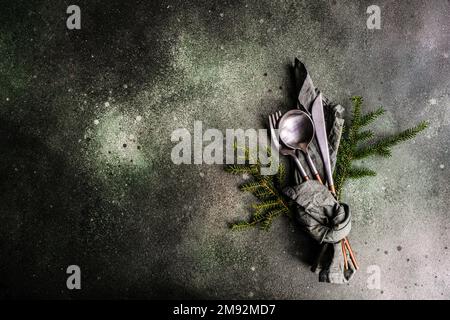 From above table setting for Christmas dinner with dark modern cutlery wrapped in green napkin with fir tree leaves and decor on concrete table Stock Photo