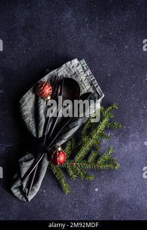 From above table setting for Christmas dinner with dark modern cutlery wrapped in green napkin with fir tree leaves and decor on concrete table Stock Photo