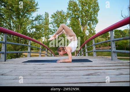 Full body side view of young flexible female practicing yoga in Vrischikasana A during outdoor workout on sunny day Stock Photo