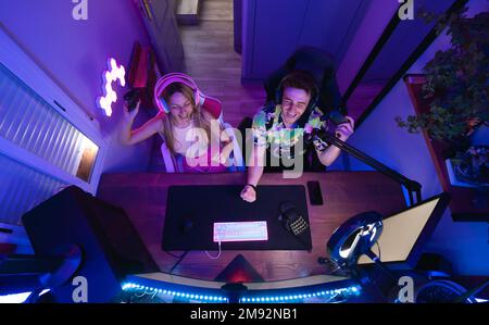 Top view of happy man and woman in wireless headphones sitting at table near camera and recording video while playing computer game Stock Photo