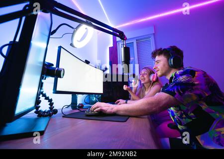 Happy man and woman in wireless headphones sitting at table near camera and recording video while playing computer game Stock Photo