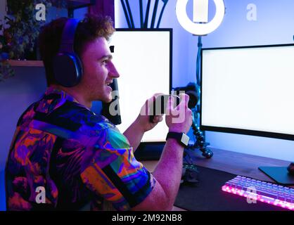 Side view of positive young male gamer in colorful shirt and wireless headset sitting near microphone while streaming and playing video game in neon i Stock Photo