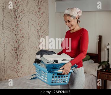 Positive mature female in casual outfit smiling and carrying blue basket with cloth after laundry routine at home Stock Photo