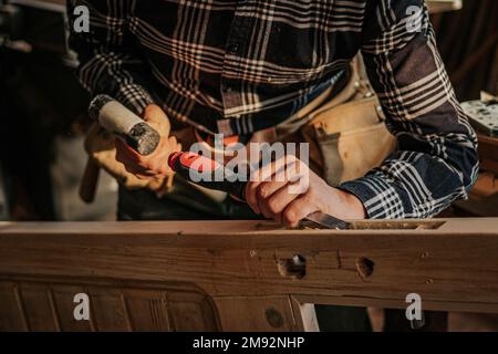 Unrecognizable male carpenter in checkered shirt cutting wooden plank with sharp chisel while working in workshop Stock Photo