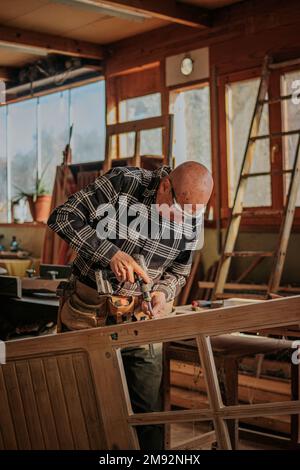 Aged male woodworker in protective glasses drilling wooden plank while working in joinery Stock Photo