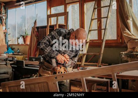 Aged male woodworker in protective glasses drilling wooden plank while working in joinery Stock Photo
