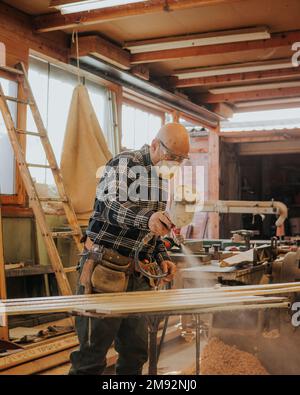 Side view of focused mature male carpenter in protective mask and goggles using electric paint sprayer while varnishing wooden plank Stock Photo