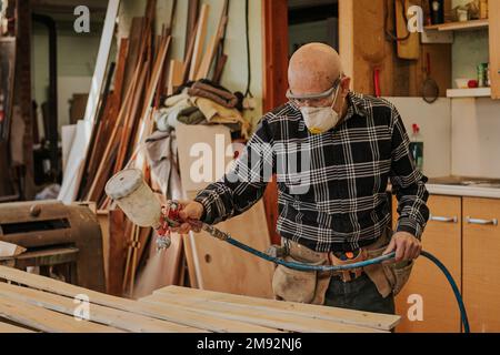 Focused mature male carpenter in protective mask and goggles using electric paint sprayer while varnishing wooden plank Stock Photo