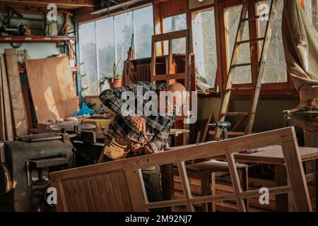 Elderly craftsman in glasses working with chisel while cutting wooden plank in workshop Stock Photo