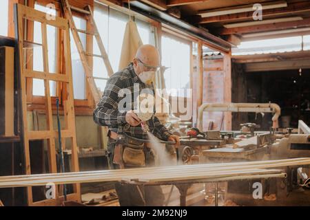 Side view of focused mature male carpenter in protective mask and goggles using electric paint sprayer while varnishing wooden plank Stock Photo