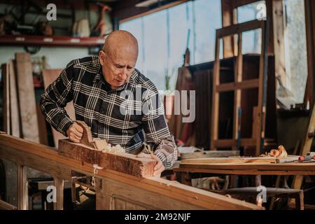 Elderly male woodworker in checkered shirt cutting wooden plank with jointer while working at workbench Stock Photo