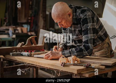Focused elderly craftsman in casual wear using ruler and pencil while measuring wooden detail in professional joinery Stock Photo