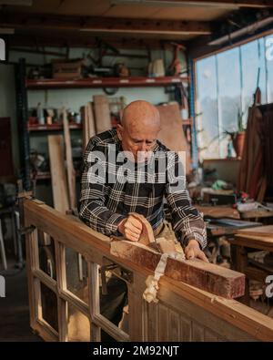 Elderly male woodworker in checkered shirt cutting wooden plank with jointer while working at workbench Stock Photo