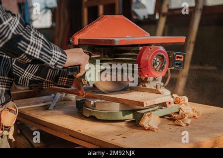 Unrecognizable male carpenter in checkered shirt using circular saw to cut wooden plank during work in joinery Stock Photo