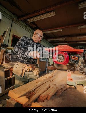 Concentrated mature male woodworker in protective goggles and headphones using circular saw to cut wooden plank during work in joinery Stock Photo