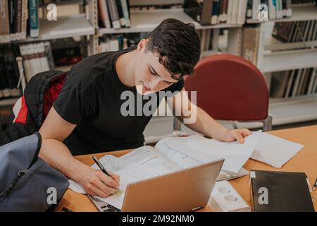 High angle of focused teen male student in casual clothes sitting at table with laptop and writing homework assignment while studying in library Stock Photo