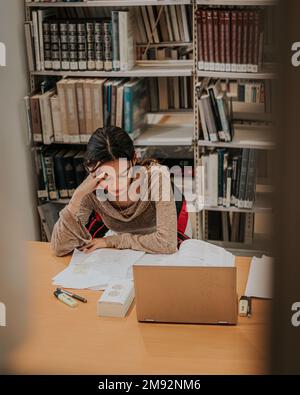 Thoughtful young female student in casual clothes sitting at wooden table with laptop while reading book during exam preparation in library Stock Photo