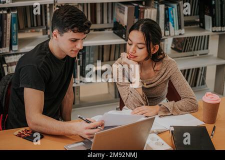 Concentrated teen students in casual outfits sitting at wooden table with laptop and working on university project together while studying in library Stock Photo