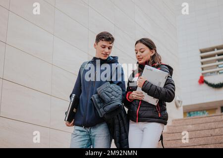 From below positive young students in warm clothes with backpacks and laptops smiling and climbing up stairs while going to university Stock Photo