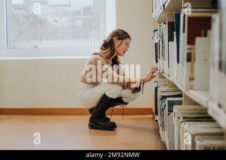 Side view full body of concentrated young Asian female student in casual clothes sitting on haunches while choosing book for reading in university lib Stock Photo