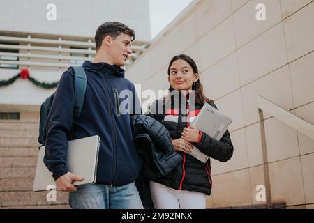 From below positive young students in warm clothes with backpacks and laptops smiling and climbing up stairs while going to university Stock Photo