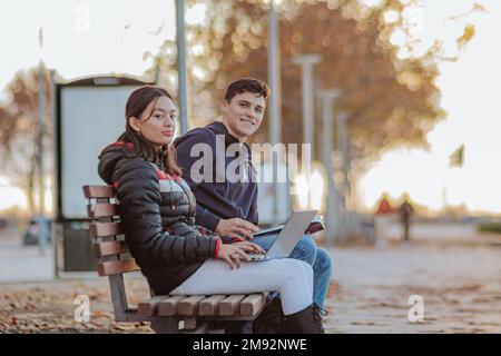 Positive multiracial friends in outerwear sitting on wooden bench and looking at camera while smiling and doing homework on laptop together in park Stock Photo