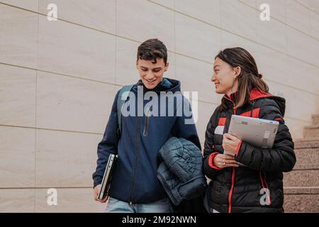 From below positive young students in warm clothes with backpacks and laptops smiling and climbing up stairs while going to university Stock Photo