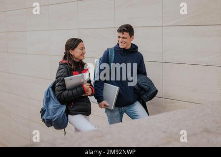 Positive young students in warm clothes with backpacks and laptops smiling and climbing up stairs looking at each other while going to university Stock Photo